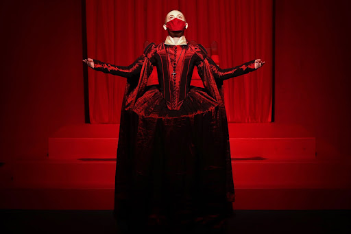a performer in a red satin dress and mask in front of a red backgroup. Their arms spread and they look up
