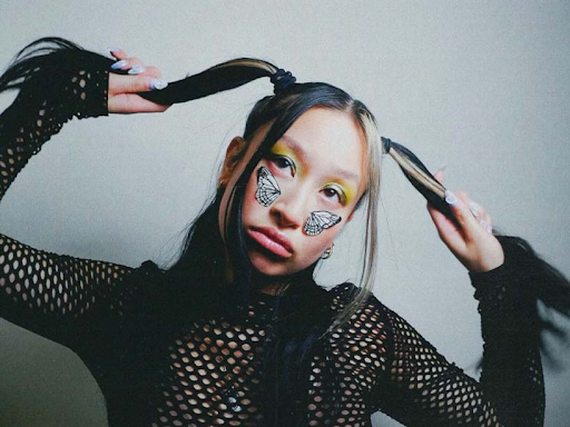 a model looks bored and pulls her pigtails to the side. She has butterfly wings on her cheeks and wears a mesh shirt