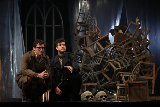 two actors crouch in front of a messy pile of chairs. They look out and talk