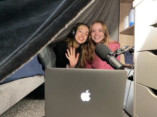 Two actors under a blanket in front of a microphone and computer smile at the camera