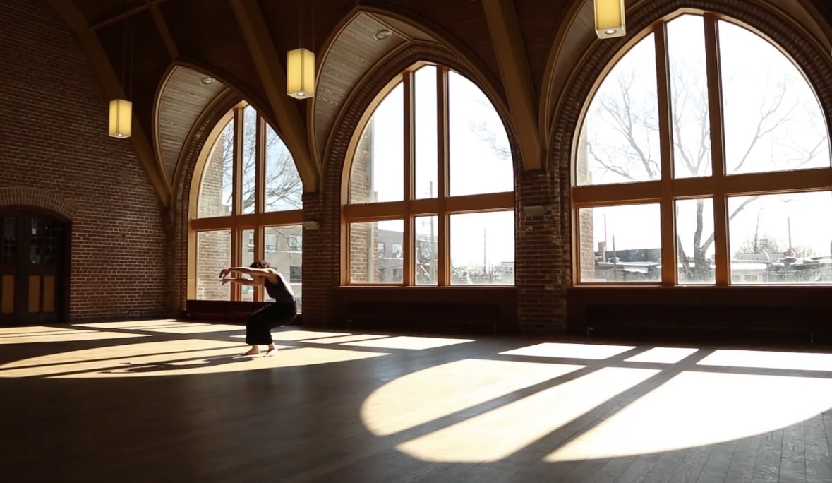 A dancer in a vast empty hall with large arched windows. The sun pours in