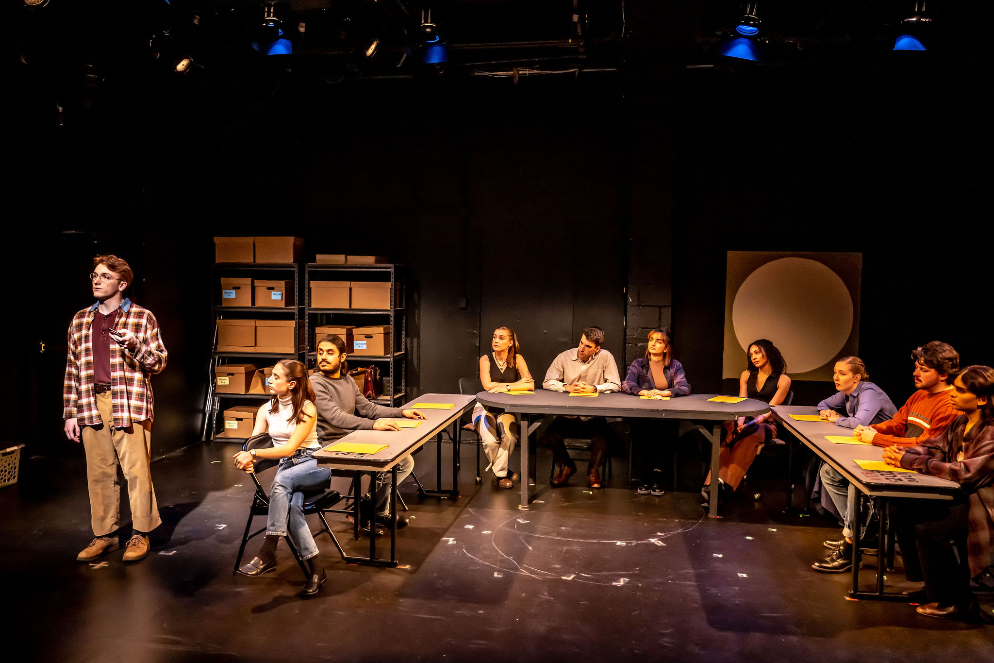 Actors sit around a table holding scripts, they are all looking at one man who is standing to the side of the tables speaking to the audience. 