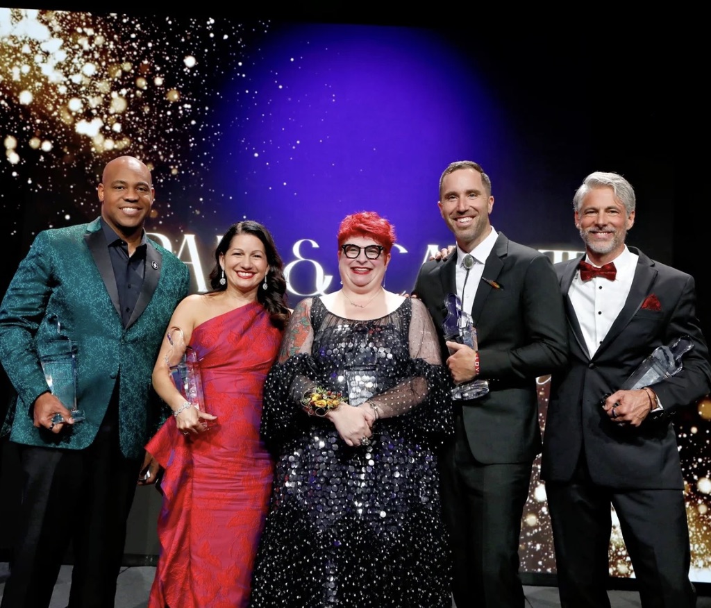 Five Individuals in formal dress stand in a line facing the camera. Each person is holding an award statue, save for the woman standing int he centre. 