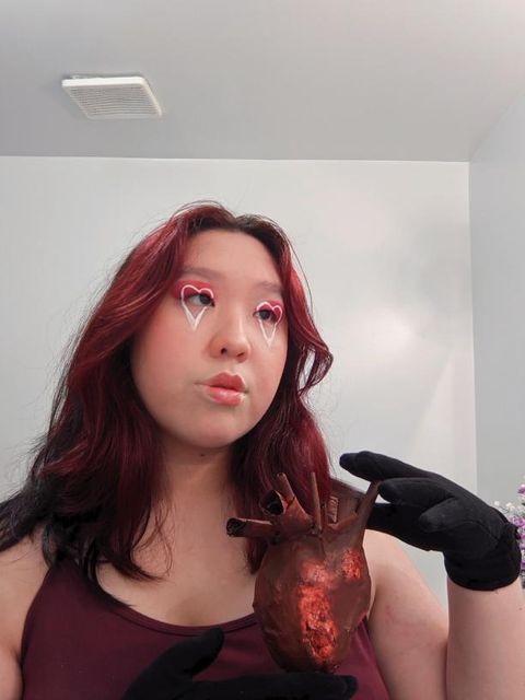 Cindy Phung, a young Asian woman  with hair died red. She has hearts painted around her eyes, and dramatic lipstick. She looks to the side and holds a papier mache heart in gloved hands