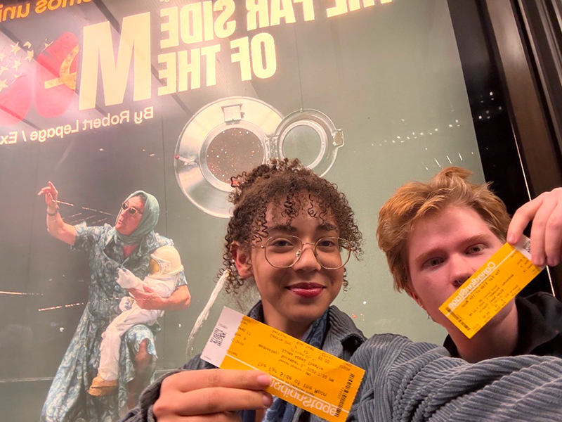  Two students pose in front of a show poster for The Other Side of the Moon at Canadian Stage, they are holding up their theatre tickets