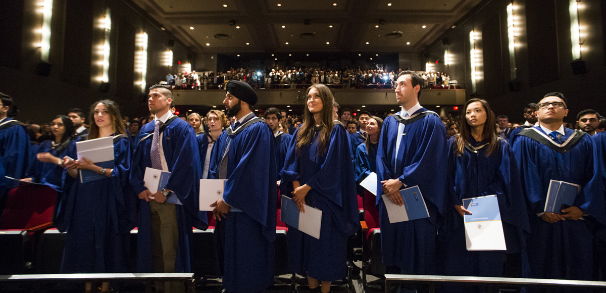 Graduates wear blue gowns and hold folders. They stand in front of their seats in the Ryerson Theatre audience.