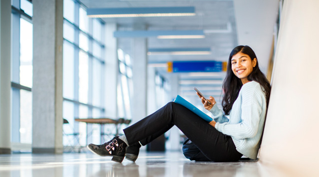 Student sitting on the floor in ENG building hallway smiling at the camera and holding a phone