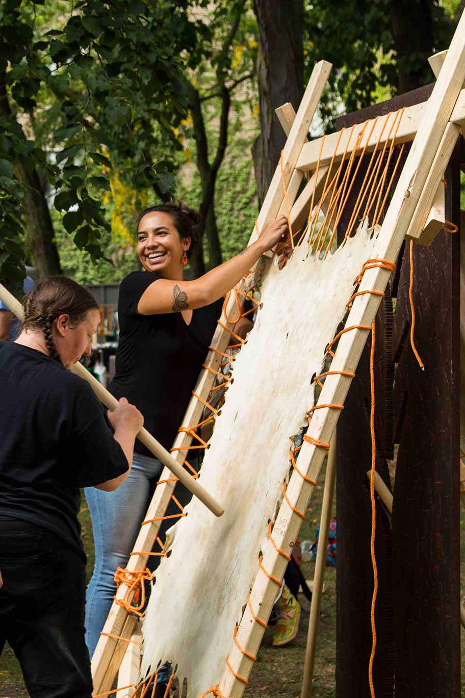 Pictured here is Amber Sandy of Scixchange, in front of the Hide Tanning at the 2019 Ryerson Pow Wow - in the Ryerson quad. The Hide is stretched over a frame, another person is working on the Hide with a large stick.