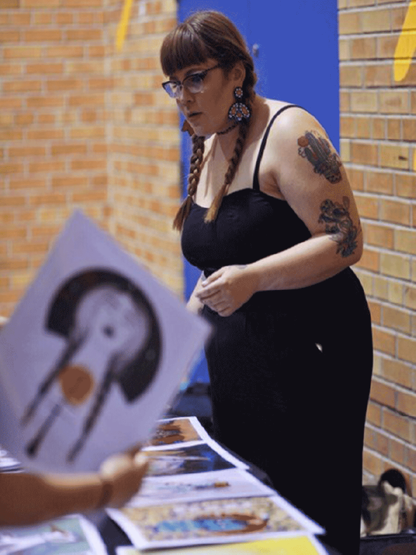 Artist Chief Ladybird sells her artwork at the 2018 Pow Wow. She leans over her table which has her various prints displayed. A customer holds up one of the prints.