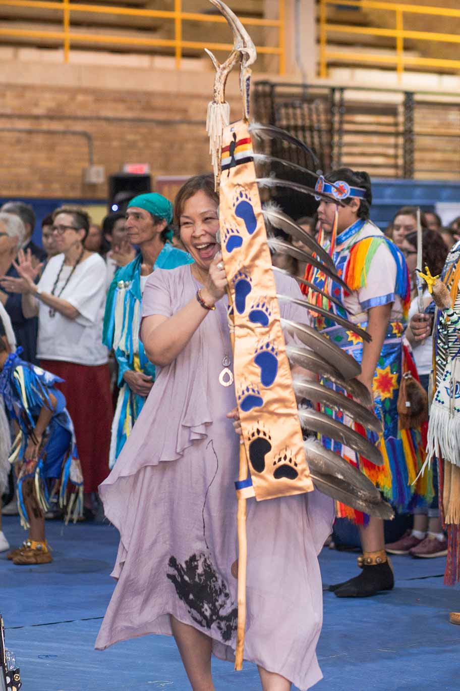 Monica McKay smiles as she carries the Ryerson Eagle Staff at the 2018 Ryerson Pow Wow. She is surrounded by Pow Wow participants in the Ryerson upper gym.