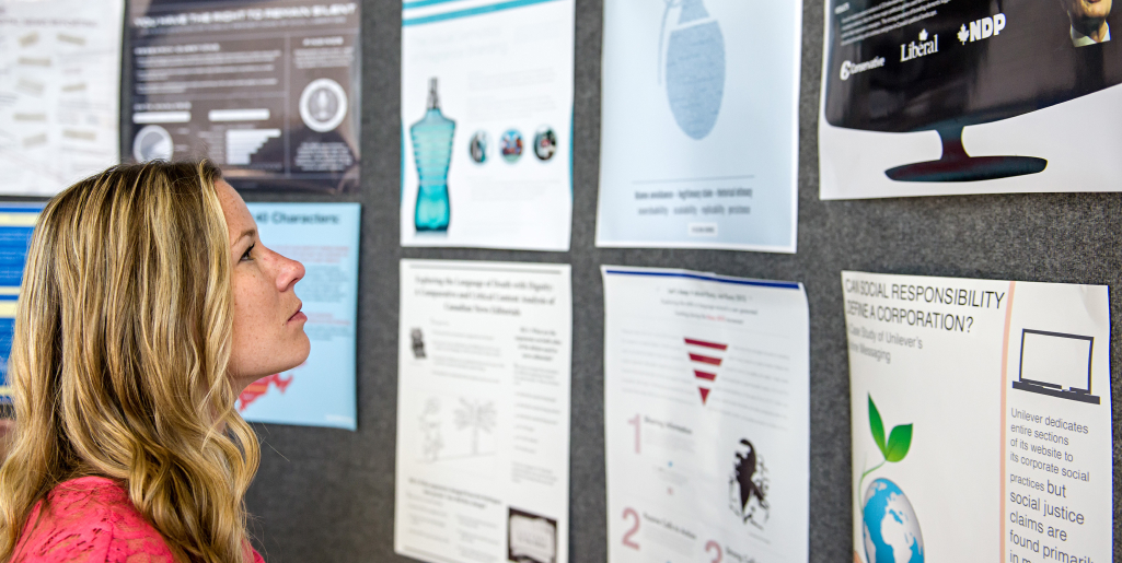 A close-up of a woman with long blonde hair wearing a bright pink top and attentively looking at a wall of academic posters displayed in a row.
