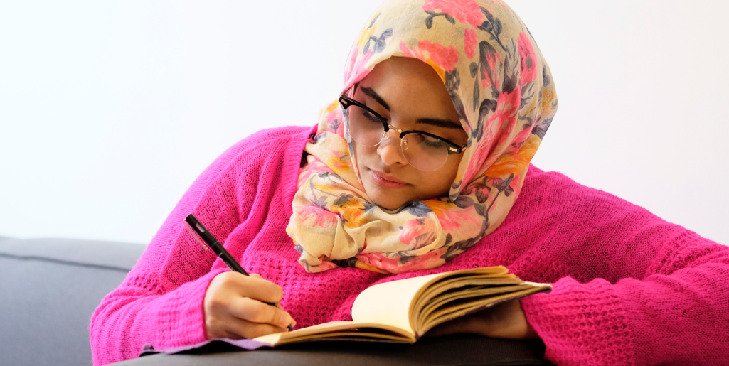 A close-up of a woman wearing glasses, a pink sweater, and a floral-patterned hijab writing in a notebook while seated and leaning forward.