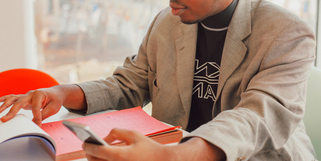 A close-up of a man wearing a grey blazer and a black graphic T-shirt flipping through a large notebook and holding a smartphone at a table.