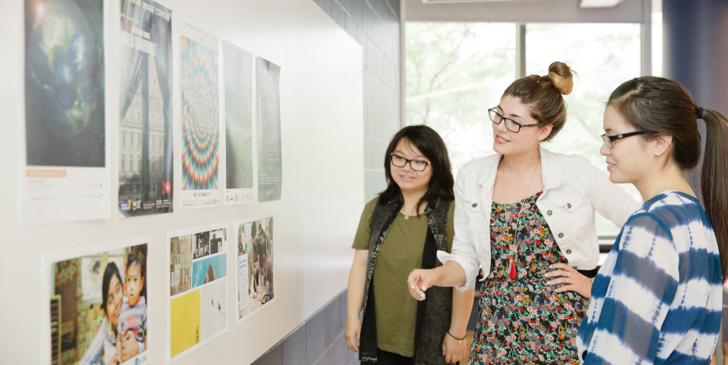 A group of three women standing in front of a wall of posters while pointing and smiling . They are discussing the displayed images. One wears a white jacket and floral dress, one wears a black jacket, and one wears a blue-and-white shirt.