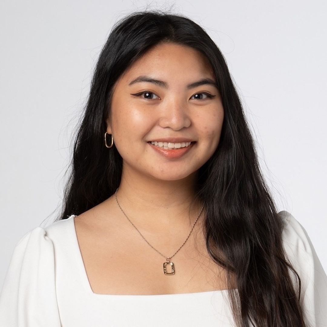 A close-up headshot of Kaitlyn Pontanilla smiling. She has long, wavy dark hair and is wearing a white square-neck blouse, small hoop earrings, and a necklace with a rectangular pendant.