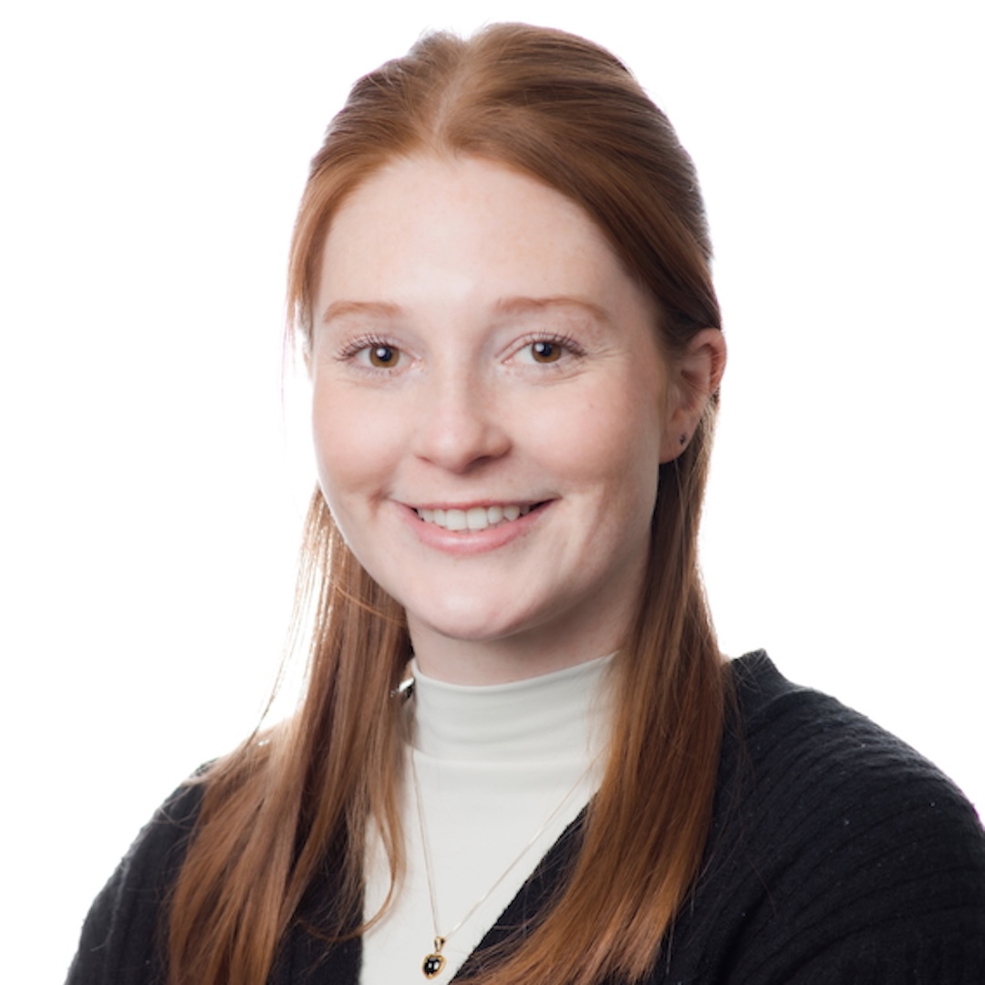 A close-up headshot of Laura Baker smiling. She has straight, long red hair and is wearing a white turtleneck, a black v-neck sweater, and a gold necklace. 