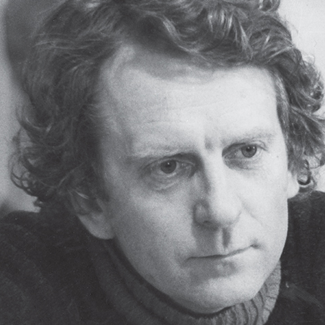 A black-and-white close-up headshot of Alan Clarke. He has short hair, glasses, and is smiling while seated at a desk in a suit and tie.