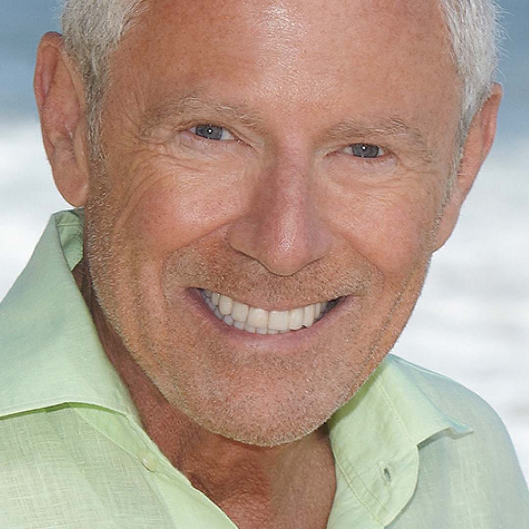 Close-up headshot of Alan Hamel smiling. He has short grey hair and is wearing a light green collared shirt with the beach in the background.