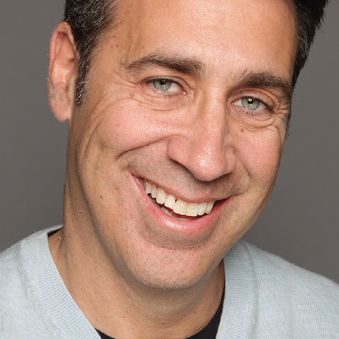 Close-up headshot of Arthur Smith smiling. He has short dark hair and is wearing a light blue sweater over a dark shirt.
