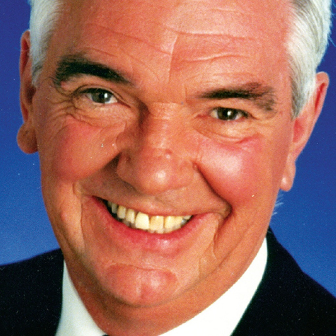Close-up headshot of Bill Lawrence smiling. He has short white hair and is wearing a dark suit with a white shirt.
