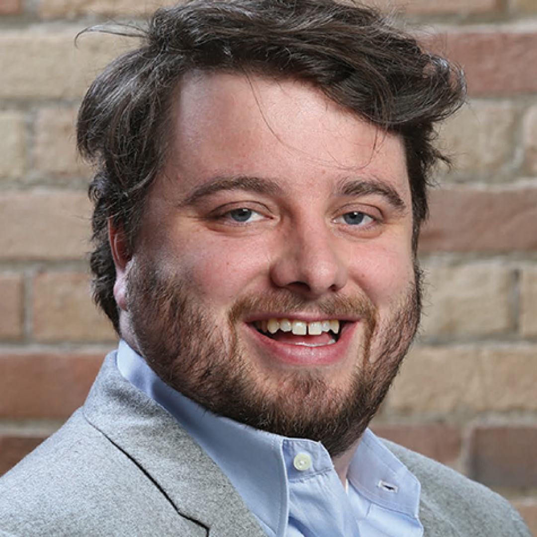 Close-up headshot of Blair Powers smiling. He has short dark hair and a beard and is wearing a light grey blazer over a collared shirt.