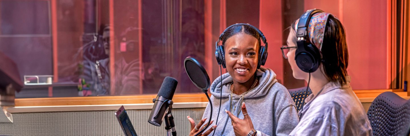 A close-up of two women wearing headphones in a recording studio. One is wearing a grey hoodie and speaking animatedly into a microphone while the other listens and smiles.
