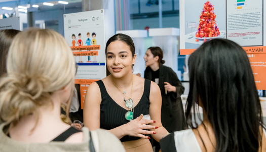 A close-up of a woman with dark hair tied back, wearing a black sleeveless top and a nametag, speaking to a small group of people in front of posters at an event.