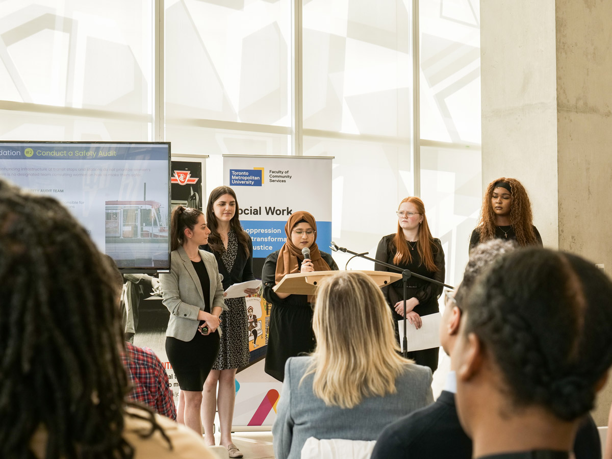 A group of students at a podium in the SLC present in front of posters for Social Work and the TTC.