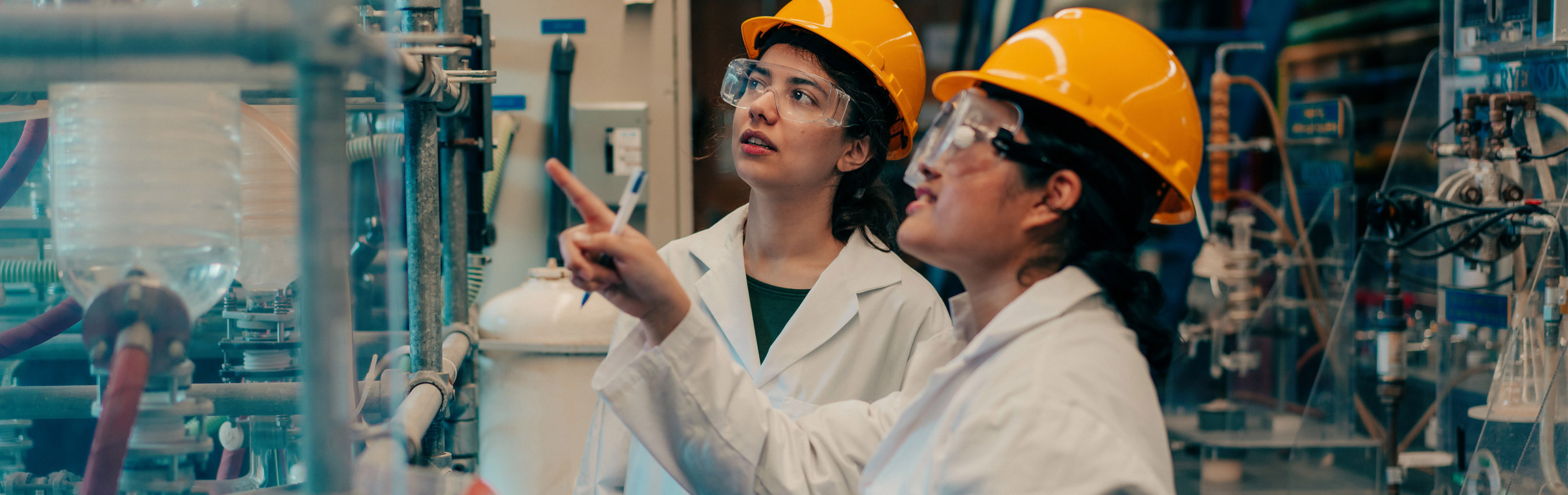 A pair of Chemical Engineering Co-op students in hard hats, goggles and lab coats discuss a project together.