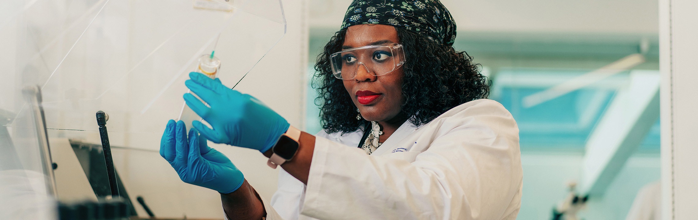 A Civil Engineering student in a lab coat, goggles and gloves inspects a project in a lab.