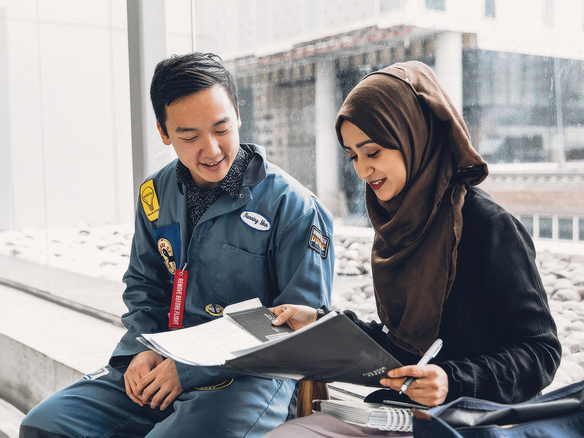 A student in TMU Engineering coveralls looks over a folder of documents with a classmate.