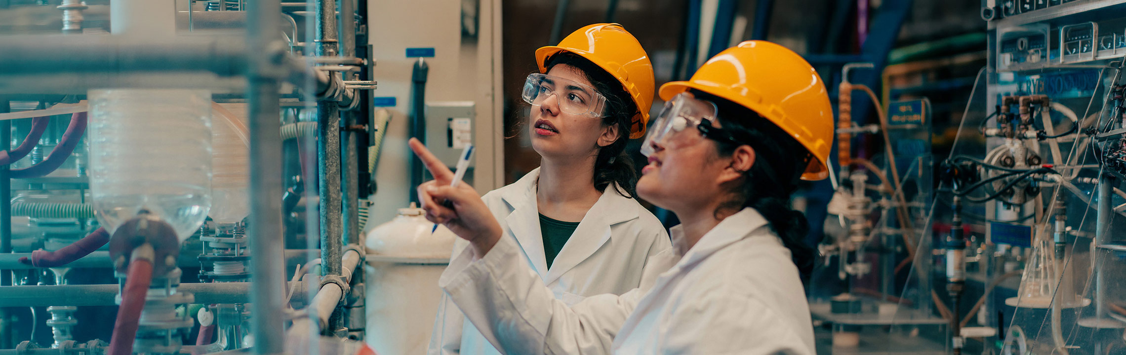 A pair of Chemical Engineering students in hard hats, goggles and lab coats discuss a project together.