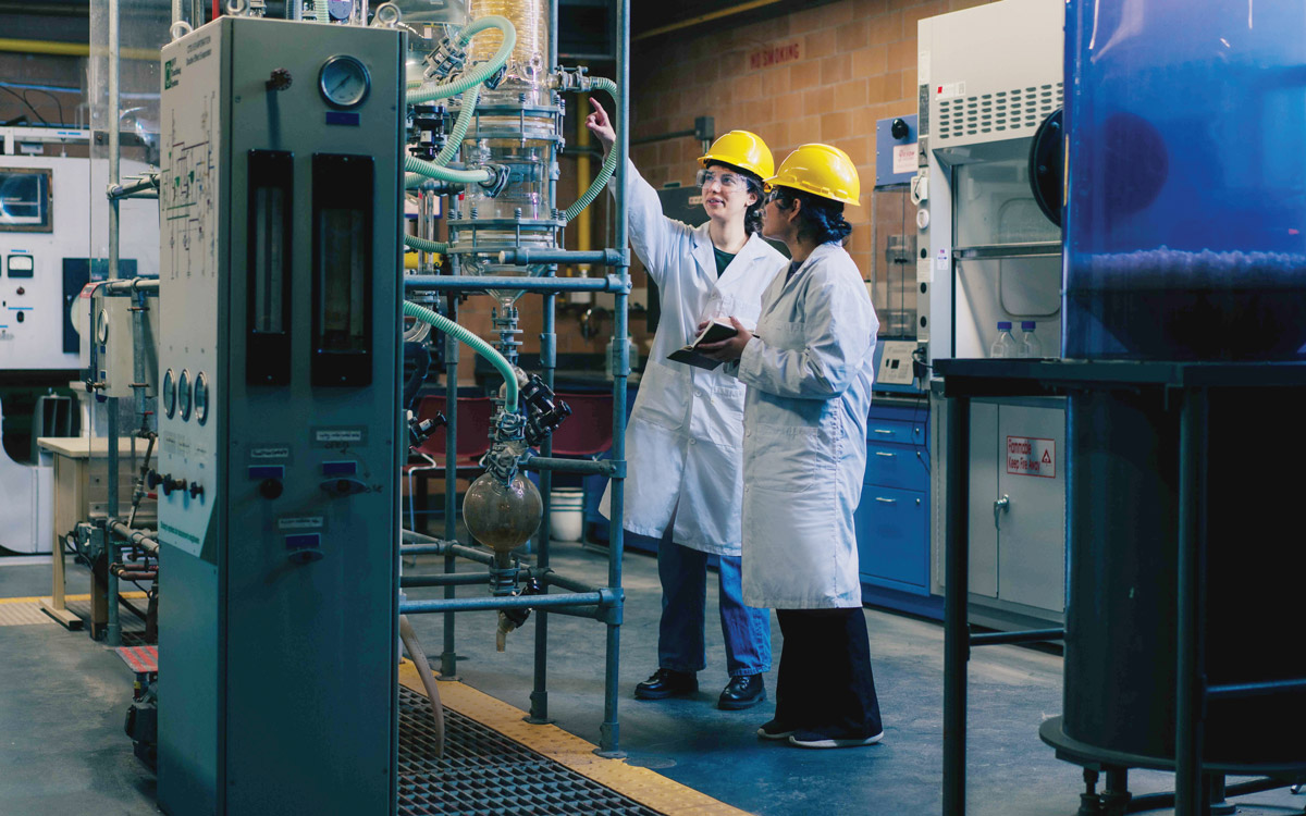 A pair of students in hard hats, goggles and lab coats discuss a project together.