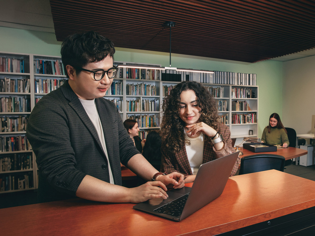 A pair of students look at a laptop screen together in the library.