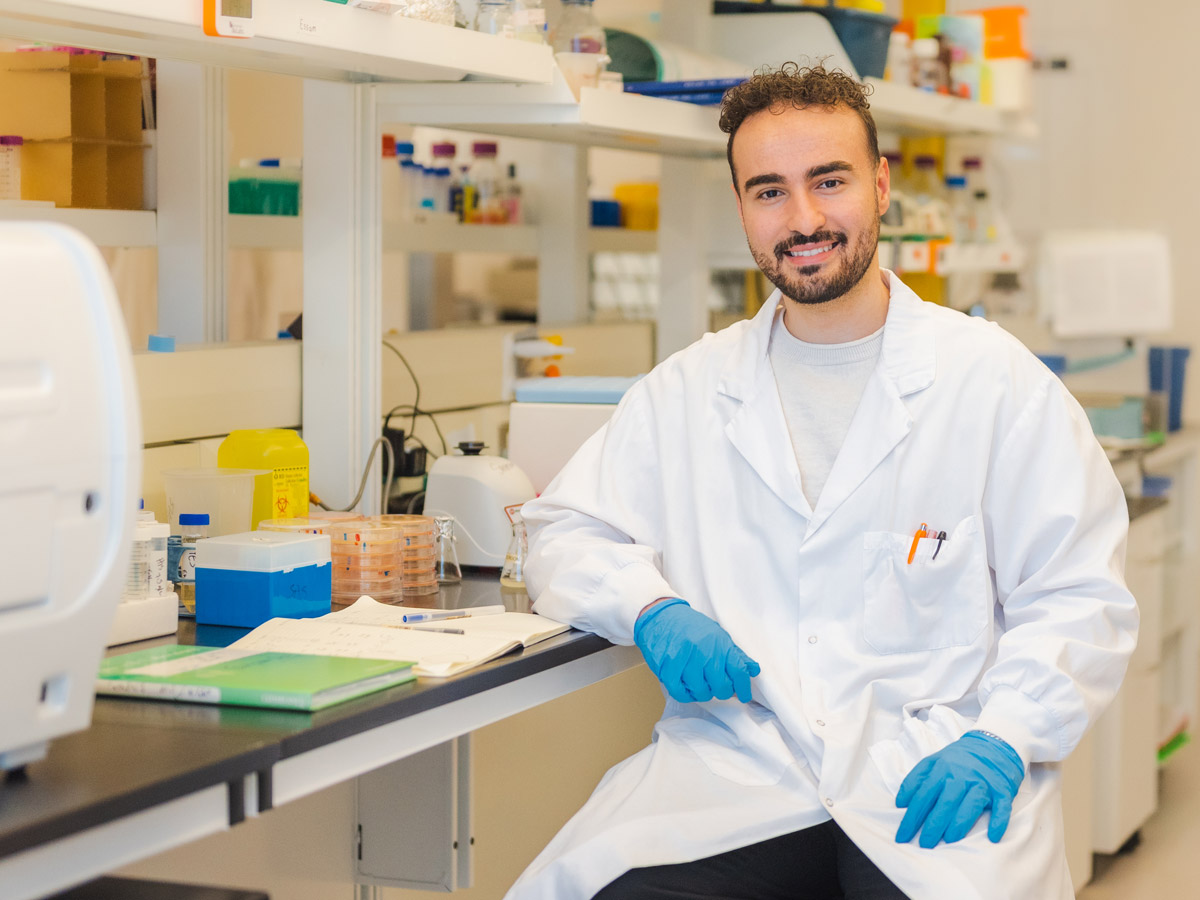 Biology graduate, Essam Karam, sitting at a lab bench wearing a lab coat and blue latex gloves.
