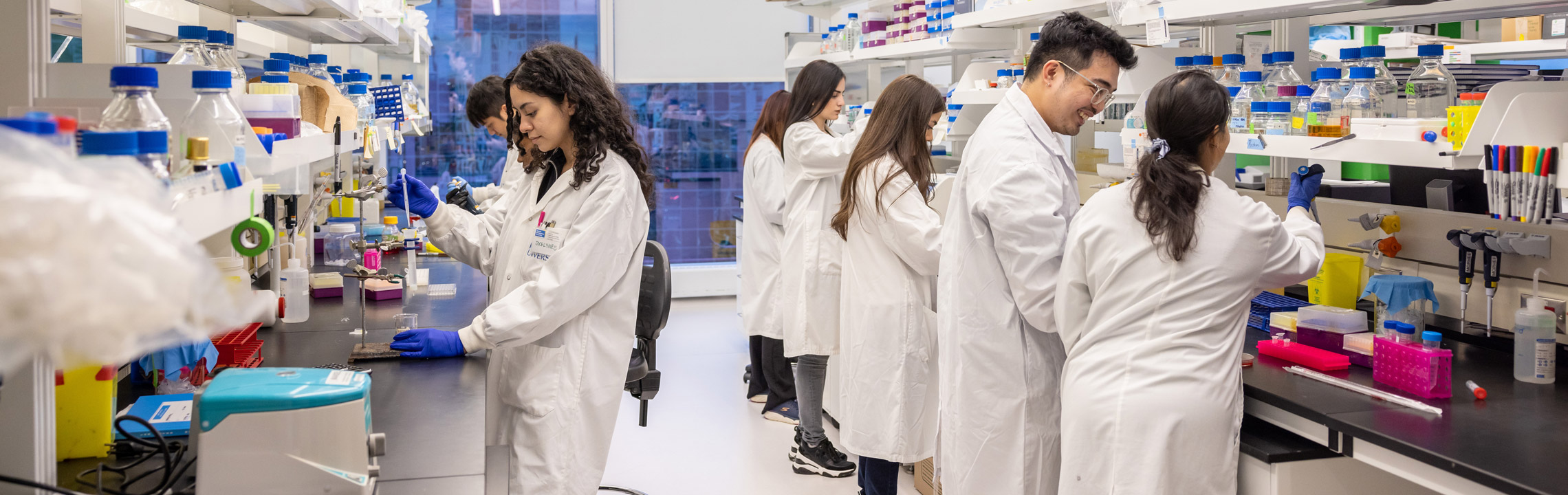 A group of Biomedical Science students in lab coats work together in a lab.