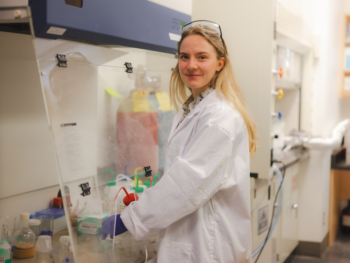 Chemistry graduate, Reese Grandy, in a lab coat, goggles and gloves working in a lab.