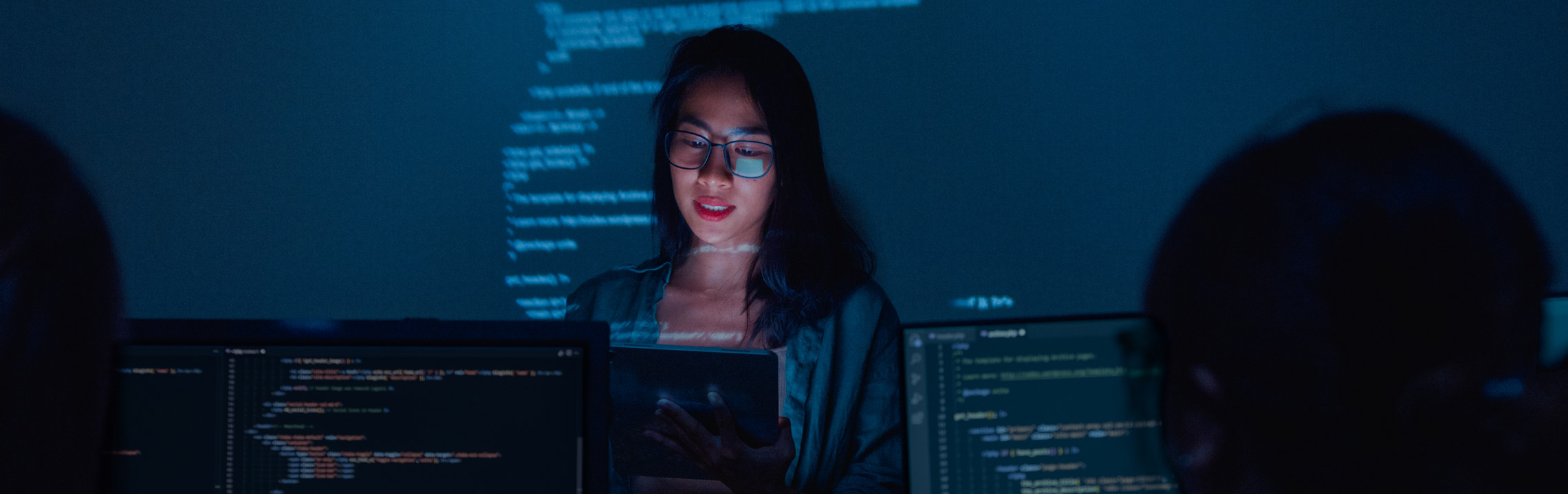A Cyber Science student, lit by the glow of a screen, presents in front of classmates in a computer lab.