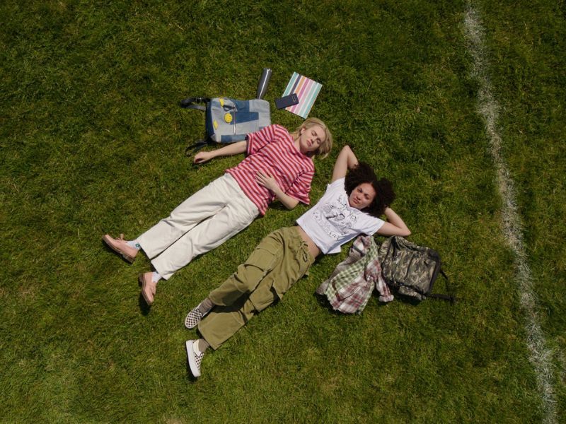 A scene from Davey & Jonesie’s Locker depicts 2 friends lying on a school field with their backpacks and belongings spread around.