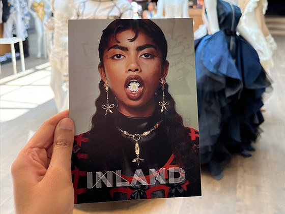 A hand holds up the flyer for INLAND. Dresses and other designs can be seen in the background.