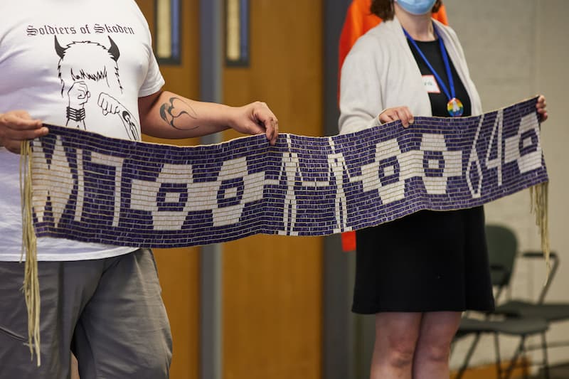 A close-up of two individuals holding a purple and white beaded wampum belt