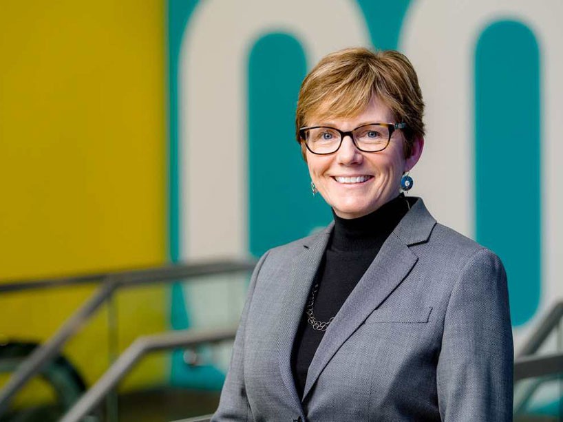 Kelly MacKay in a grey suit with black shirt in front of a colourful teal and yellow wall