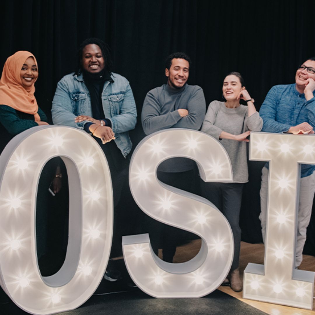 Five students from diverse backgrounds smile and lean against large letters O,S, I that are lit up.