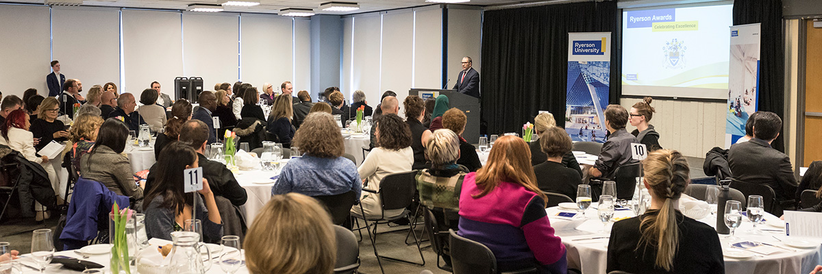 A large crowd shot of tables of people sitting looking at the front of the room and a speaker in front of a screen.