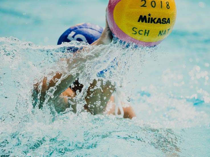 A Water Polo player in the water obscured by water splashes, wearing a blue cap and holds a yellow ball in one hand.  