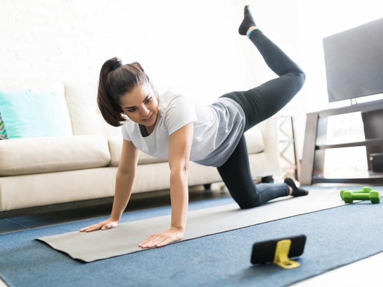 A female is performing donkey kicks in her living room while following a virtual fitness class on her phone.