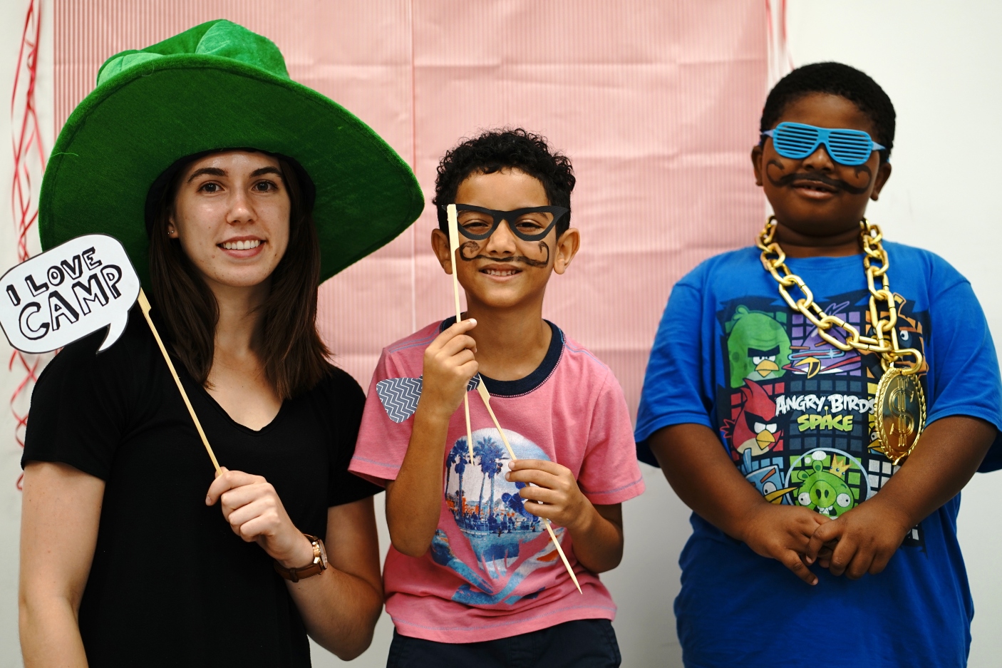 A camp counsellor and two campers wearing dress-up hats and glasses. The counsellor holds a sign that read, "I love camp."