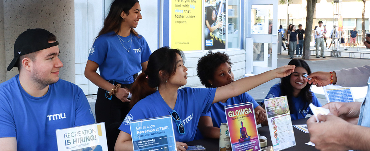 Student staff in blue shirts sit at a table providing information to students asking about recreation programs.