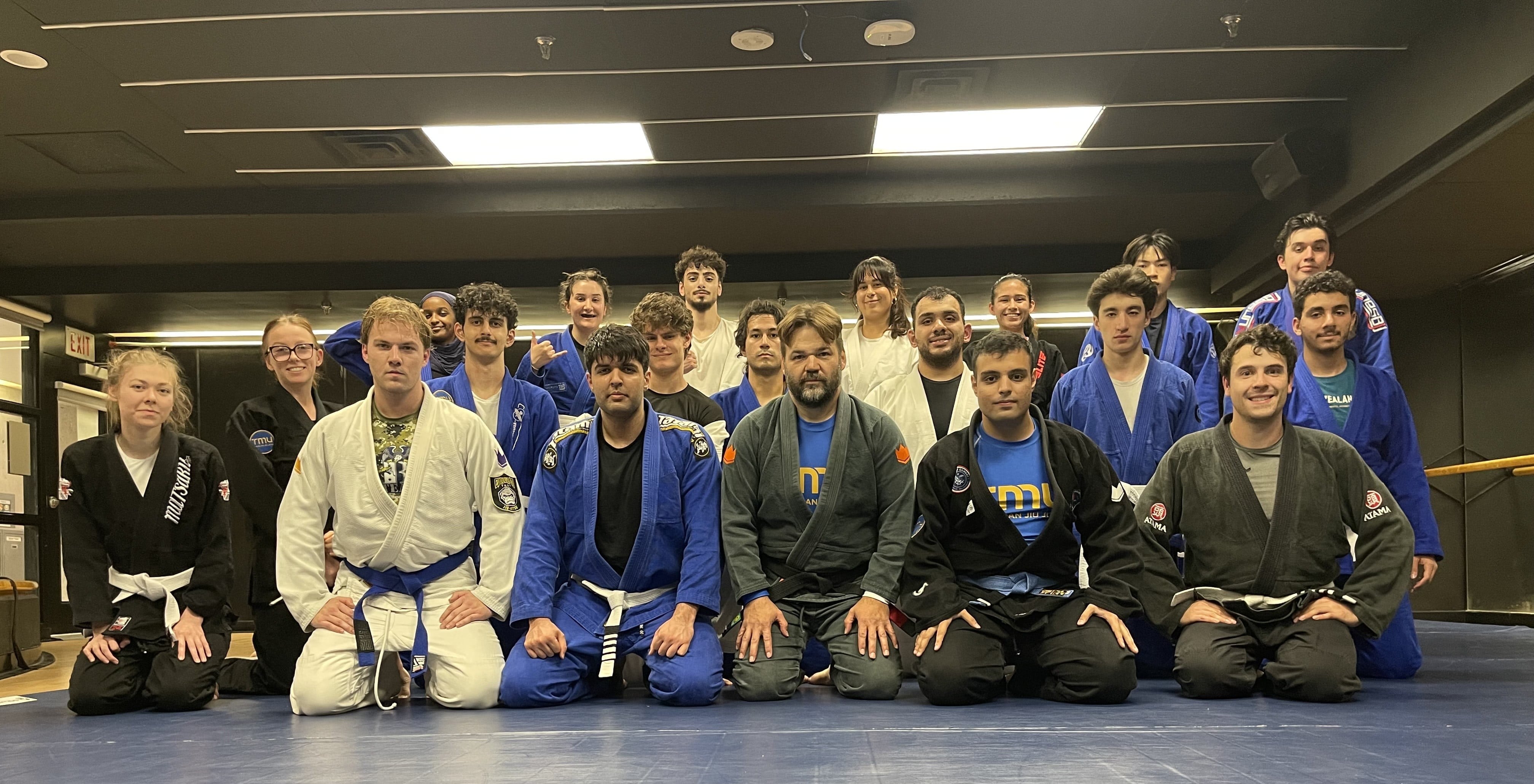 Two men practice Brazilian Jiu Jitsu on a blue mat surrounded by their class.