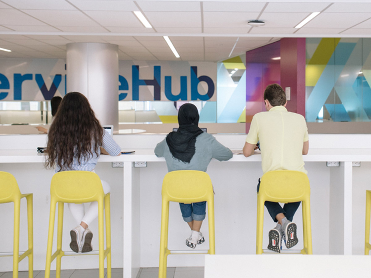 Three students sit on the yellow chairs across from the ServiceHub in the Podium building.